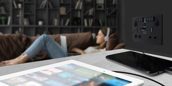 Lady using a laptop wearing headphones in the background in the foreground are 2 devices on charge from BG Electrical evolve decorative light switches and sockets