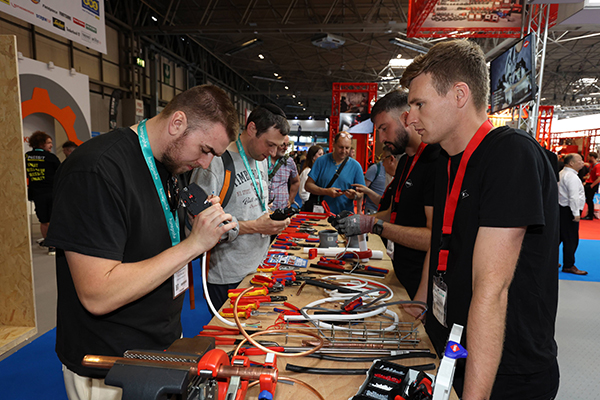 InstallerSHOW 6 people stood around a large table full of electrical gadgets, tools and wires, inlcuding a clamp housing a piece copper pipe.
