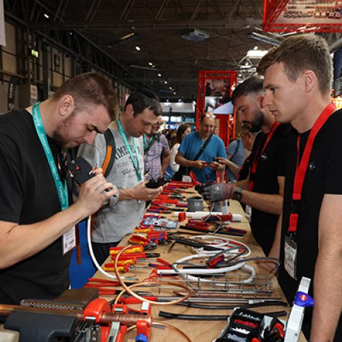 InstallerSHOW 6 people stood around a large table full of electrical gadgets, tools and wires, inlcuding a clamp housing a piece copper pipe.